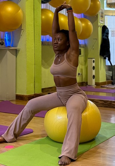 Mujer con ropa deportiva beige estira los brazos por encima de la cabeza mientras está sentada en una pelota amarilla de ejercicio en un gimnasio con paredes verdes y esterillas de yoga.