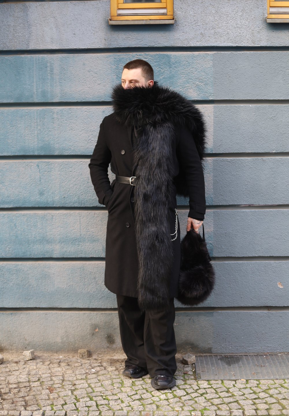 Manteau noir avec un col en fourrure texturée et un long accent en fourrure, associé à un pantalon noir et un sac à main en fourrure, debout devant un mur bleu.