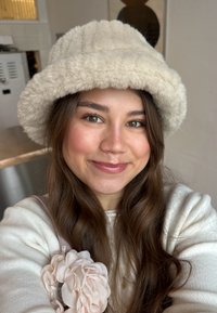 Jeune femme aux longs cheveux bruns portant un chapeau seau beige duveteux et un pull blanc, souriant à l'intérieur sous une lumière naturelle douce.
