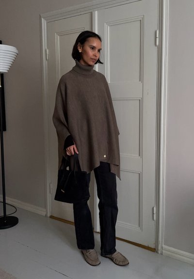 Mujer con un poncho de cuello alto marrón, pantalones negros y mocasines beige, sosteniendo un bolso negro, de pie en el interior junto a una puerta blanca.