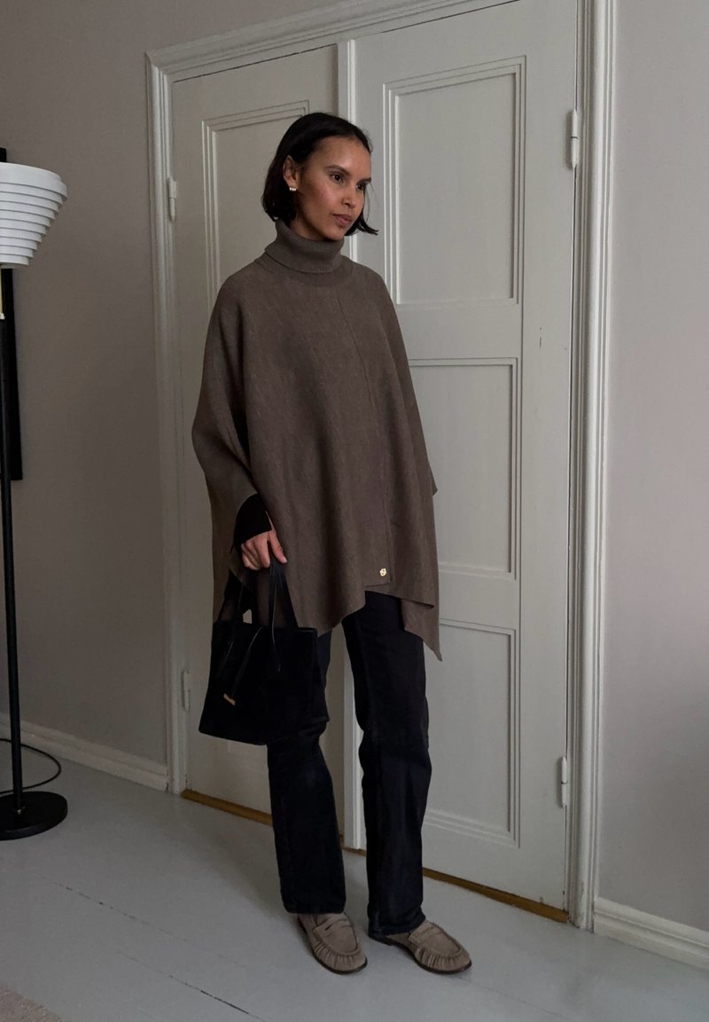 Femme portant un poncho à col roulé marron, pantalon noir et mocassins beiges, tenant un sac à main noir, debout à l'intérieur près d'une porte blanche.