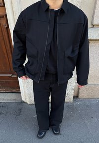 Personne portant une tenue entièrement noire comprenant une veste, un pull, un pantalon large et des chaussures, debout sur un trottoir en ville près d'une porte en bois.