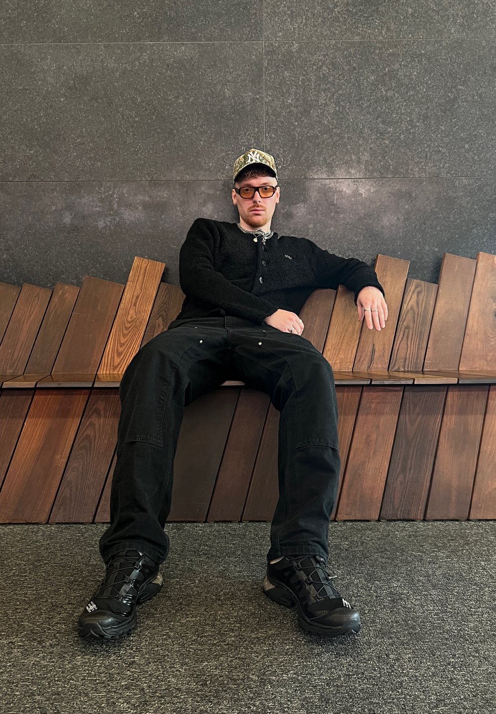 Cardigan en polaire noire boutonné en haut, pantalon cargo noir et baskets noires. Assis sur un banc en bois au design à lattes.