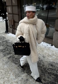 Femme marchant sur un trottoir enneigé portant un manteau blanc en fausse fourrure, un béret blanc, un pantalon blanc, des gants noirs, des chaussures noires pointues, et portant un sac à main noir.