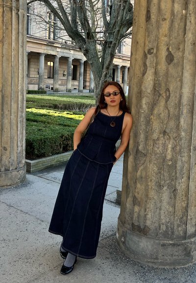 Joven mujer con vestido oscuro y gafas de sol apoyada contra una gran columna de piedra, con un edificio histórico y un jardín al fondo.