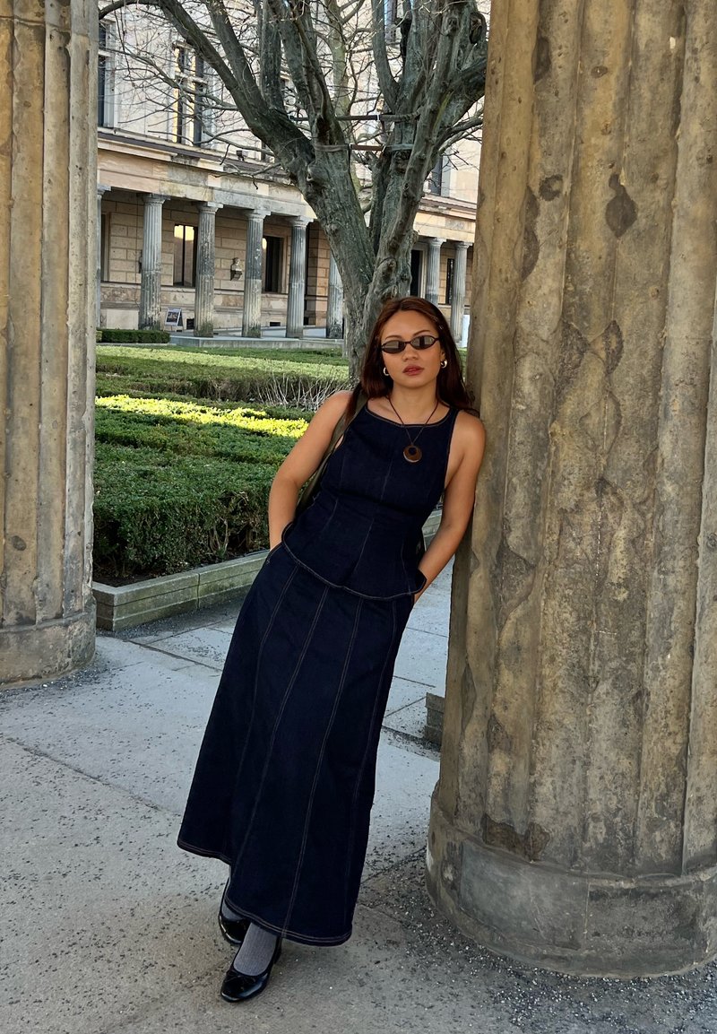 Jeune femme en robe foncée et lunettes de soleil appuyée contre une grande colonne en pierre, avec un bâtiment historique et un jardin en arrière-plan.