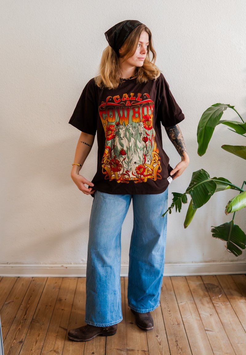 Ung kvinde iført en sort t-shirt med grafisk tryk "Cowboy", vidde jeans, et bandana og støvler, stående ved siden af en stor grøn plante indendørs.