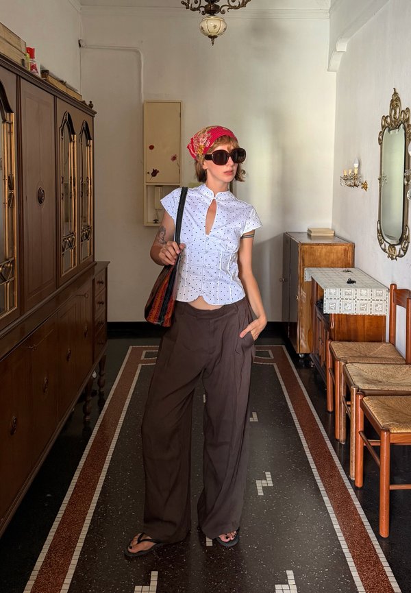 Giovane donna con occhiali da sole, foulard rosso, camicetta bianca a pois e pantaloni larghi scuri, ferma in una stanza vintage con mobili in legno.