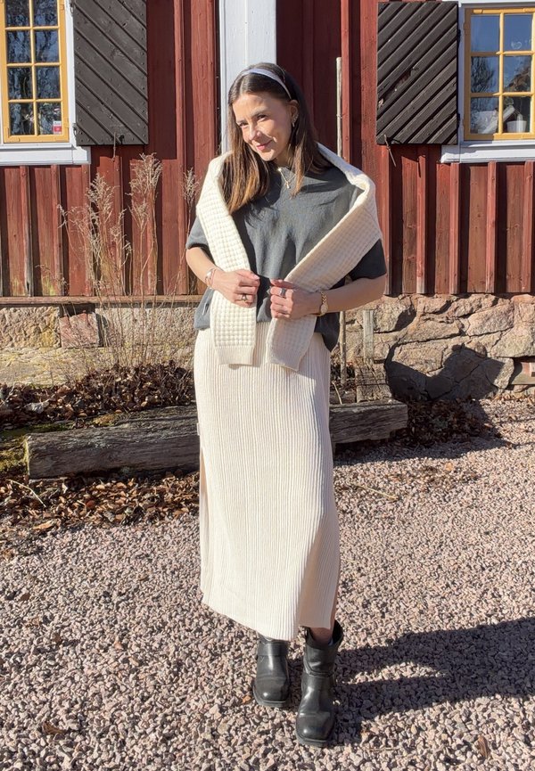 Woman standing outdoors in front of a red wooden building, wearing a gray top, cream knit skirt, cream sweater draped over shoulders, and black boots.