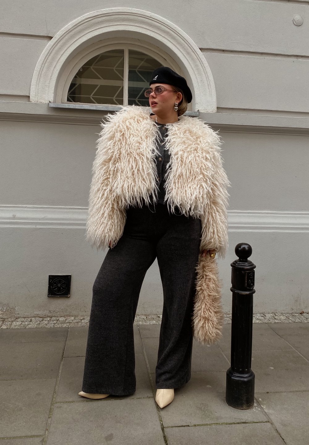 Ψεύτικο γούνινο μπουφάν σε κρέμα με χνουδωτή υφή, μαύρη μπερέ, γκρι παντελόνια φαρδιάς γραμμής και ανοιχτόχρωμες μυτερές γόβες, αξεσουάρ με μια φουντωτή τσάντα.