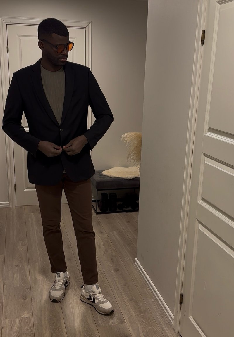 Homme portant des lunettes de soleil, un blazer noir, un pantalon marron et des baskets blanches, debout à l'intérieur en boutonnant son blazer.