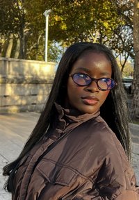 Brun pufferjakke med høy krage, med en frontlomme. Runde, lilla tonede briller. Langt, rett hår. Myk utendørsbelysning.