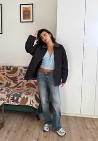 Donna con capelli scuri che indossa una giacca nera, un crop top azzurro chiaro, jeans e sneakers, in piedi all'interno accanto a un divano a fantasia e un armadio bianco.