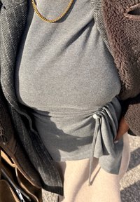 Gros plan d'une personne portant une robe grise avec un cordon de serrage latéral, un manteau en chevrons gris, une veste polaire marron, un collier chaîne en or et une jupe beige.