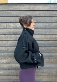 Veste noire courte en tissu épais, avec un col montant, des boutons décoratifs sur les manches et un motif texturé. Portée par-dessus une robe violette.