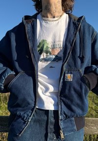 Kék farmerdzseki cipzárral, bordázott mandzsettákkal és Carhartt logóval. Alatta egy fehér grafikai póló látható, amelyen autó és fák vannak.
