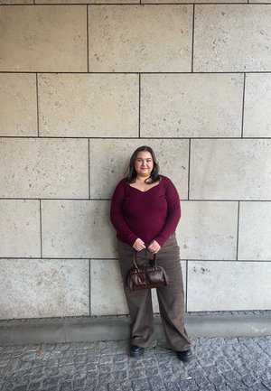 Top burdeos de manga larga acanalado, pantalones marrones de pana de pierna ancha y un pequeño bolso marrón. Pared de piedra beige texturizada al fondo.