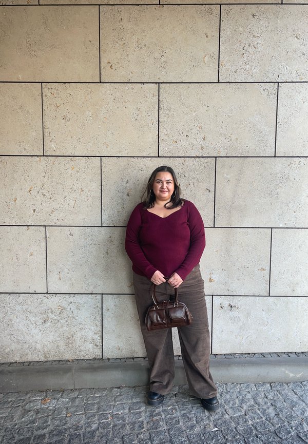 Vino rdeča rebrasta majica z dolgimi rokavi, rjave široke kordurove hlače in majhna rjava torbica. V ozadju teksturirana bež kamnita stena.