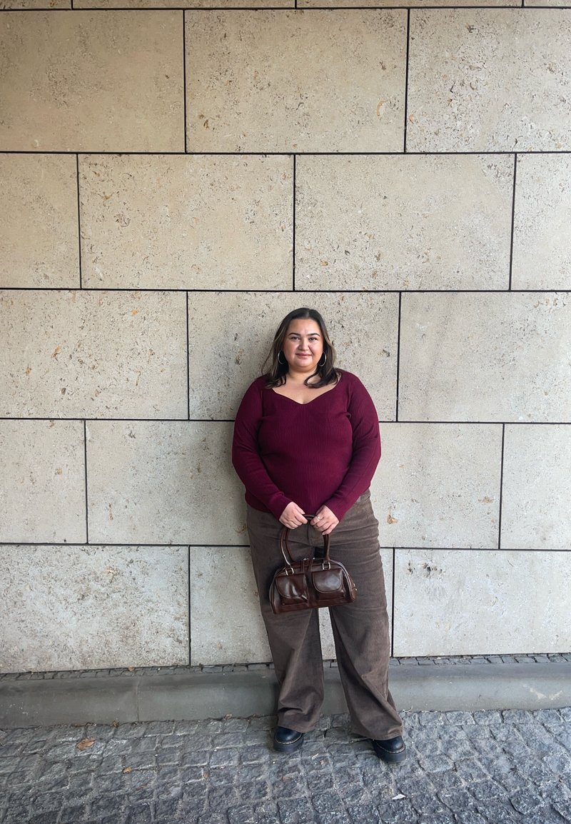 Burgundy ribbed long-sleeve top, brown wide-leg corduroy trousers, and a small brown handbag. Textured beige stone wall in the background.