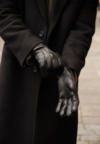 Gants en cuir noir avec des accents cousus, portés par une personne vêtue d'un manteau noir. Les gants présentent une surface texturée et un ajustement serré.