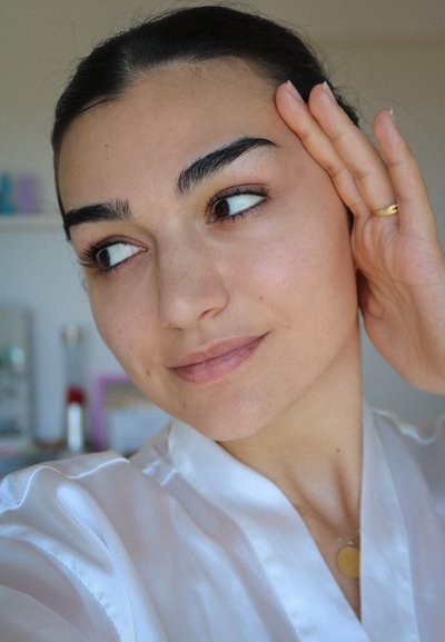 Bata de satén blanca con un collar dorado redondo. La persona tiene el cabello oscuro, piel suave y se está tocando la cara, mirando de lado.