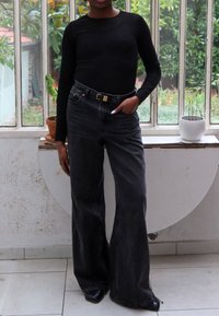Persona de pie en interiores, vistiendo una camiseta de manga larga negra con acanalado, pantalones negros de tiro alto y pernera ancha, y zapatos de punta negra.