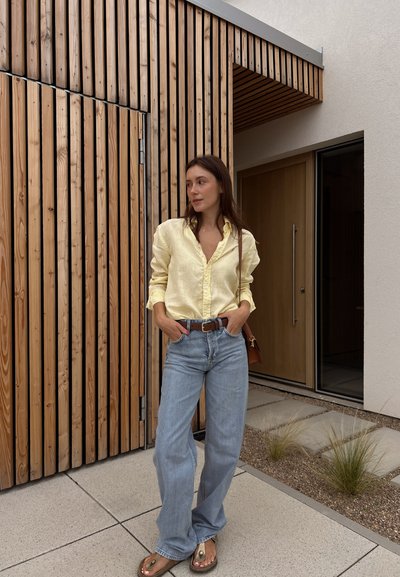 Camisa de lino amarillo claro con un corte relajado, jeans de cintura alta azul claro, cinturón de cuero marrón y sandalias de goma, en un entorno arquitectónico moderno.