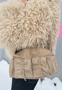 Sac à bandoulière en suède beige avec poches avant froncées, porté avec un pantalon noir et une veste texturée crème duveteuse.