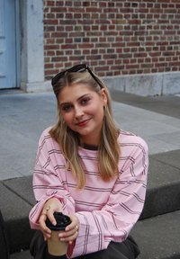 Rosa gestreifter Pullover mit langen Ärmeln, braunen Akzenten, hält eine schwarze Kaffeetasse, sitzt auf Betonstufen vor einer Backsteinmauer.