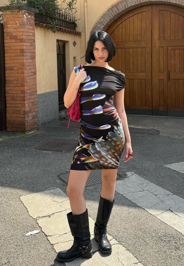 Kobieta z krótkimi czarnymi włosami w kolorowej sukience w kwiaty, czarnych butach i czerwoną torebką stoi na przejściu dla pieszych przed drewnianymi drzwiami.