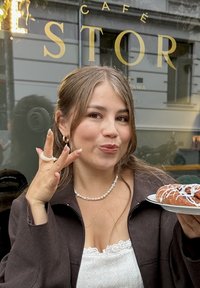 Un croissant au chocolat sur une assiette blanche tenu par une personne portant une veste marron foncé. L'individu porte un collier de perles et des bagues, montrant un geste ludique.
