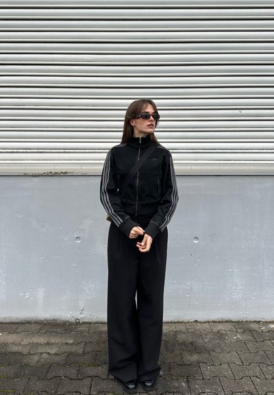 Mujer con chaqueta deportiva negra y pantalones anchos, usando gafas de sol, de pie sobre un suelo pavimentado contra una pared gris con una persiana metálica arriba.