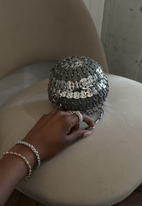Hand mit Silber- und Goldarmbändern sowie Ringen, die auf einem beigen Stuhl ruhen, neben einer runden Metallhandtasche mit verknüpften Silberstücken.