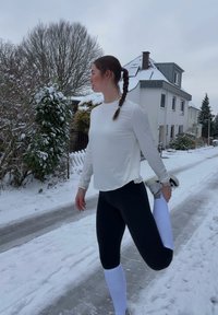 Chemise blanche à manches longues et leggings noirs, avec des jambières blanches texturées. Rue enneigée et verdure en arrière-plan.