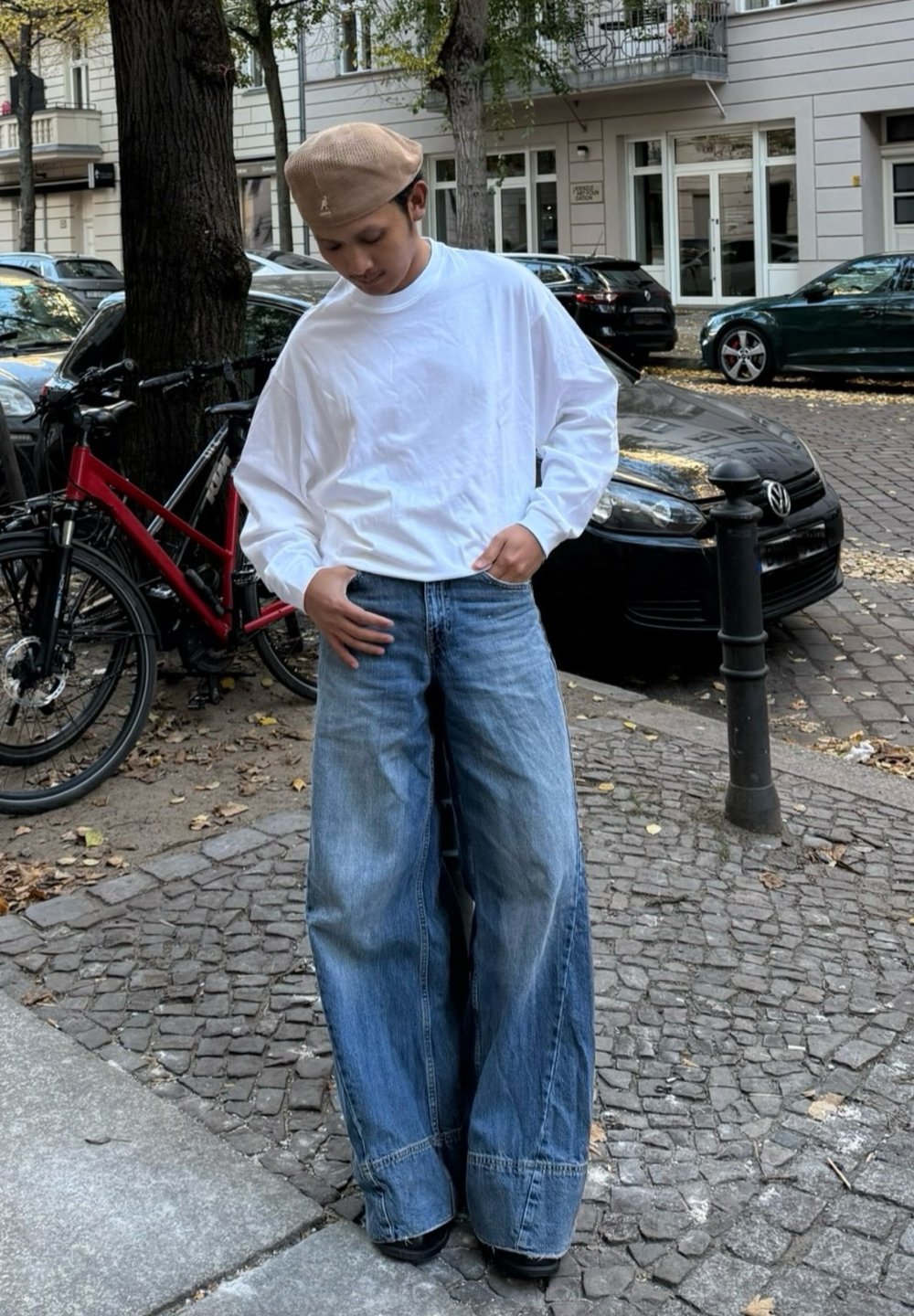 Weit geschnittene blaue Jeans mit ausgewaschenem Effekt, kombiniert mit einem locker sitzenden weißen Langarmshirt und einem hellbeigen Barett. Urbane Umgebung.