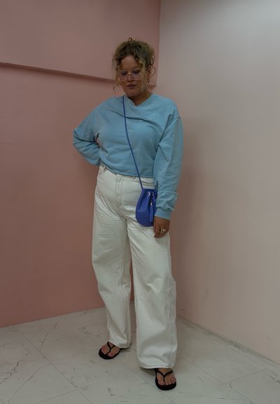 Sudadera azul claro, jeans de pierna ancha blancos, bolso cruzado morado, chanclas negras. Cabello rizado, de pie contra una pared rosa.