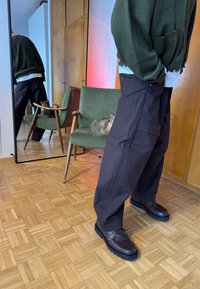 Personne portant un pull vert foncé et un pantalon noir ample se tenant près d'une chaise verte avec un chat, sol en bois et mur miroir en arrière-plan.