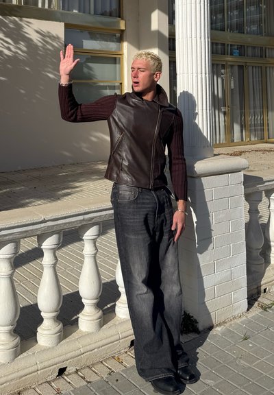 Chaleco de cuero marrón sobre una camiseta de punto de manga larga oscura, combinado con unos vaqueros oscuros sueltos y zapatos negros, frente a un fondo de columnas claras.