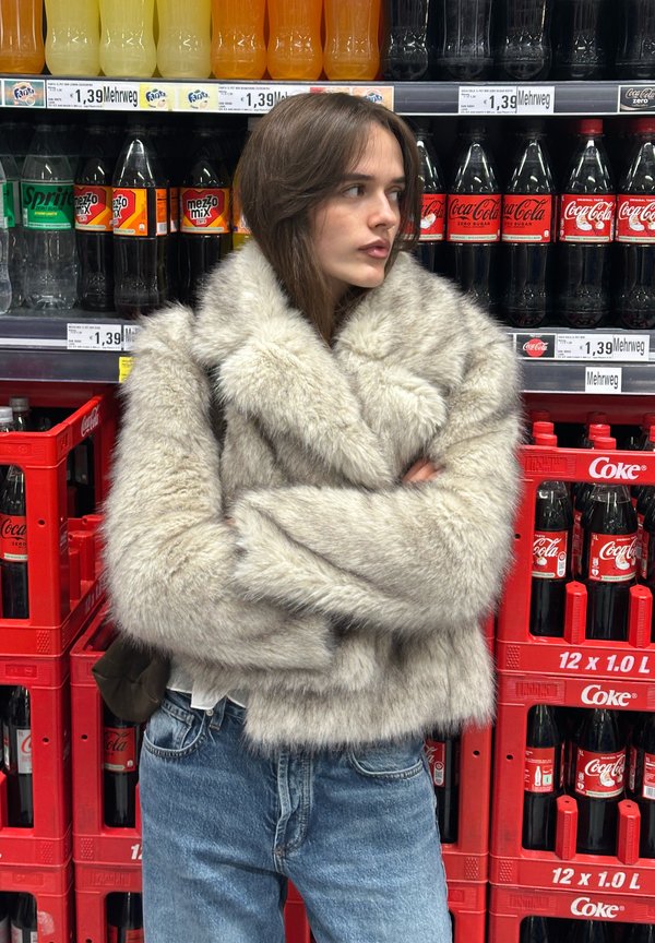 Szürke műszőrme kabát magas gallérral és széles ujjakkal, fehér ing és farmer fölött viselve, italpolcok hátterével.