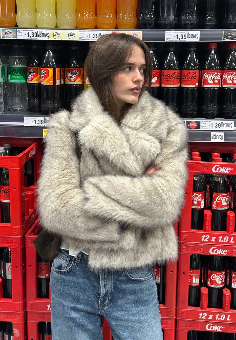 Pilko netikrų kailių paltas su aukštu apykakliu ir plačiomis rankovėmis, dėvimas ant baltos palaidinės ir džinsų fono su gėrimų lentynomis.