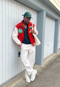 Casaco varsity vermelho e creme com emblemas, camisa preta de botões, calças brancas de perna larga e sapatilhas altas. Um boné verde completa o visual.
