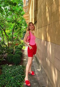 Rosa ribbet topp, røde shorts med lommer, røde sandaler med sløyfedetaljer, og en rosa mønstrete skulderveske. Omgitt av grønt.