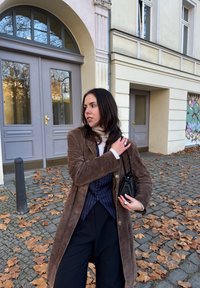 Brūns zamšāda mētelis, svītrains vestes tops, melnas augstā jostasvieta bikses, tur rokā mazu melnu somiņu. Āra vidē, uz zemes krīt lapas, fonā redzama ēka.