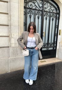 Beige rutigt kavaj, vit linne, högmidjade blå jeans, vita sneakers och en krämfärgad väska med en dekorativ stängning. Svart järngunga i bakgrunden.