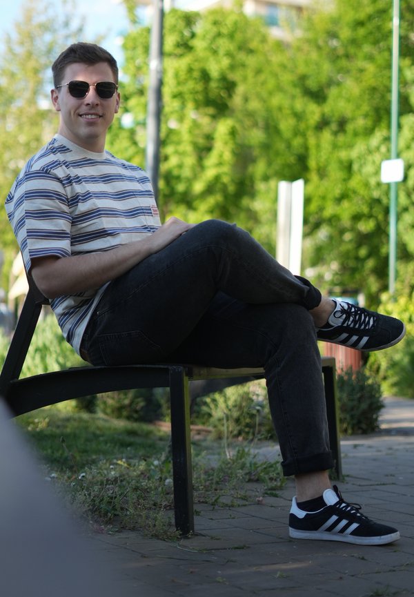 Fiatal férfi napszemüvegben és csíkos pólóban keresztbe tett lábbal ül egy parkban egy padon, háttérben zöld fák.