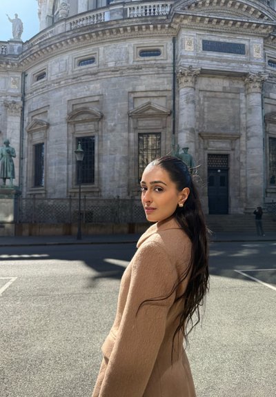 Abrigo de lana de color beige claro con cuello de chale. La modelo tiene el cabello largo y oscuro, está de pie contra un edificio histórico de piedra, con una luz suave que proyecta sombras.