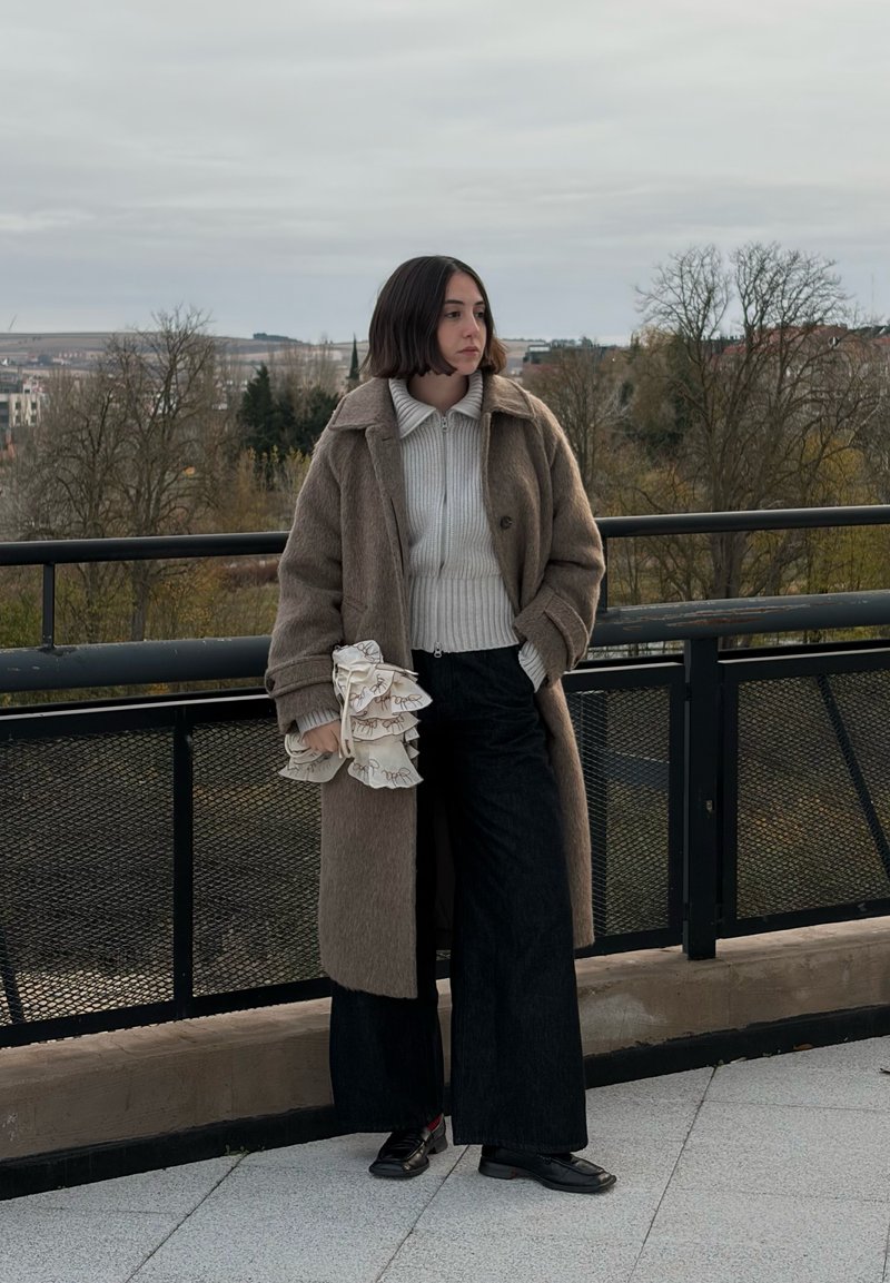 Brown wool coat over a grey knitted jumper and black wide-leg trousers; holding a ruffled white bag. Outdoor setting with trees in the background.