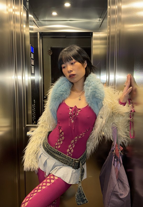 Jeune femme en tenue rose à lacets, veste blanche duveteuse avec col bleu, ceinture en jean et jupe à motifs posant à l'intérieur d'un ascenseur en métal.