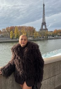 Jeune femme portant un grand manteau en fourrure noire appuyée contre une balustrade en pierre près de la Seine, avec la tour Eiffel et des arbres d'automne en arrière-plan.