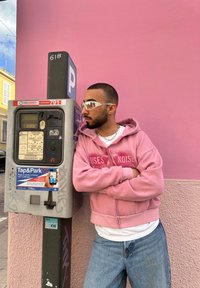 Parkirna ura z sivo kovinsko ohišje, digitalnim zaslonom in modrim nalepko Tap&Park. Ozadje sestavlja enotna roza stena. Oseba v roza kapuci in sončnimi očali.